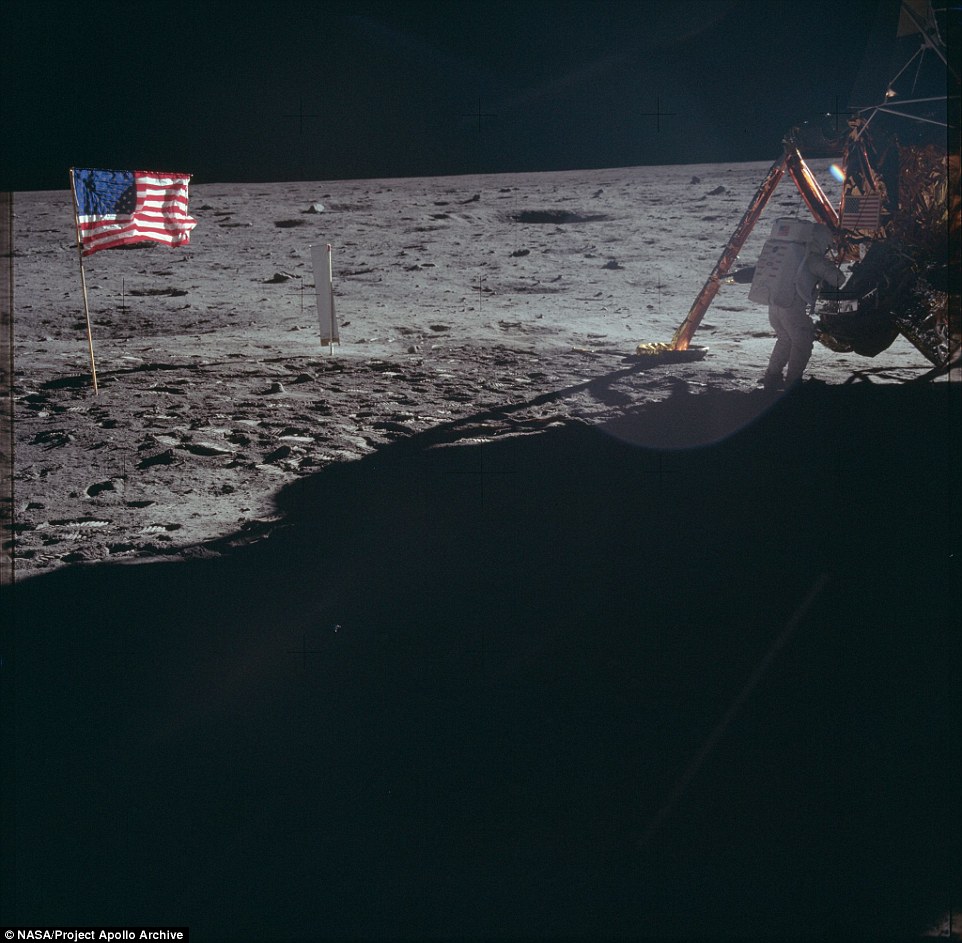 One of the astronauts fiddles with something in the Lunar Module while an American flag is planted behind him 